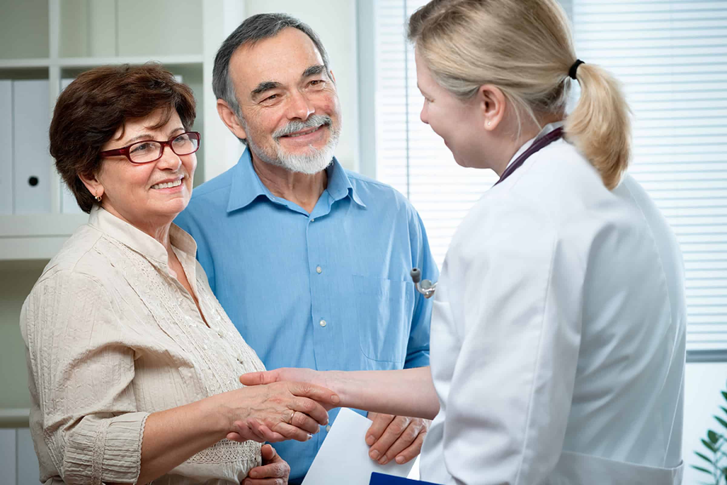 Healthcare professional shakes hands with an older couple during a consultation in a medical office.