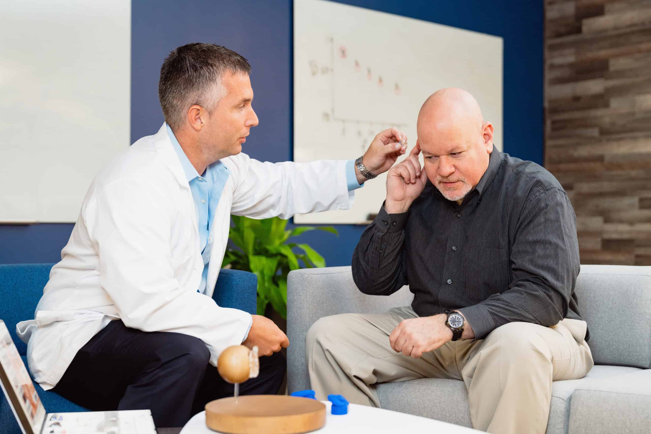 Hearing aid specialist in white coat assisting senior male patient with inserting hearing aid while seated on gray couch in comfortable office setting. The patient concentrates as he places the device in his ear with guidance from the specialist. Modern office features blue accent wall, anatomical ear chart, green plant, and educational materials on coffee table, creating a welcoming environment for hearing care.