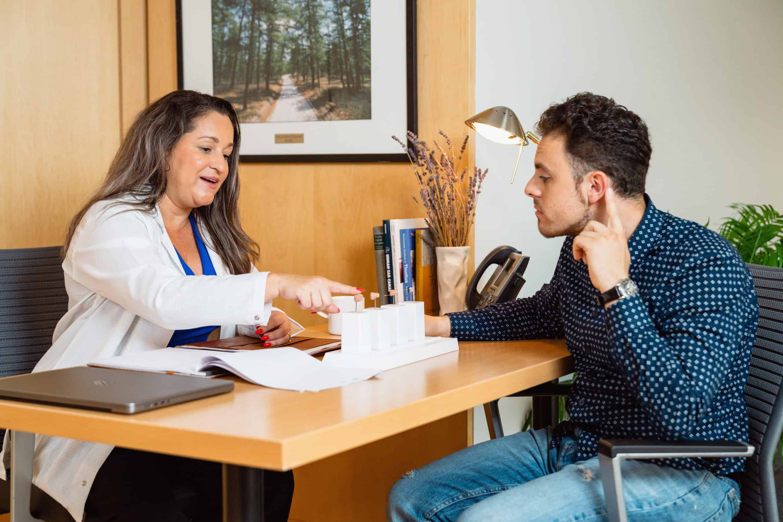 Hearing aid specialist in white coat showing hearing aid models and documentation to male patient across desk in professional office. The specialist gestures toward hearing aid samples in a display case while the attentive patient listens. Warm office setting features framed forest photography, desk lamp, books, and laptop on wooden desk.