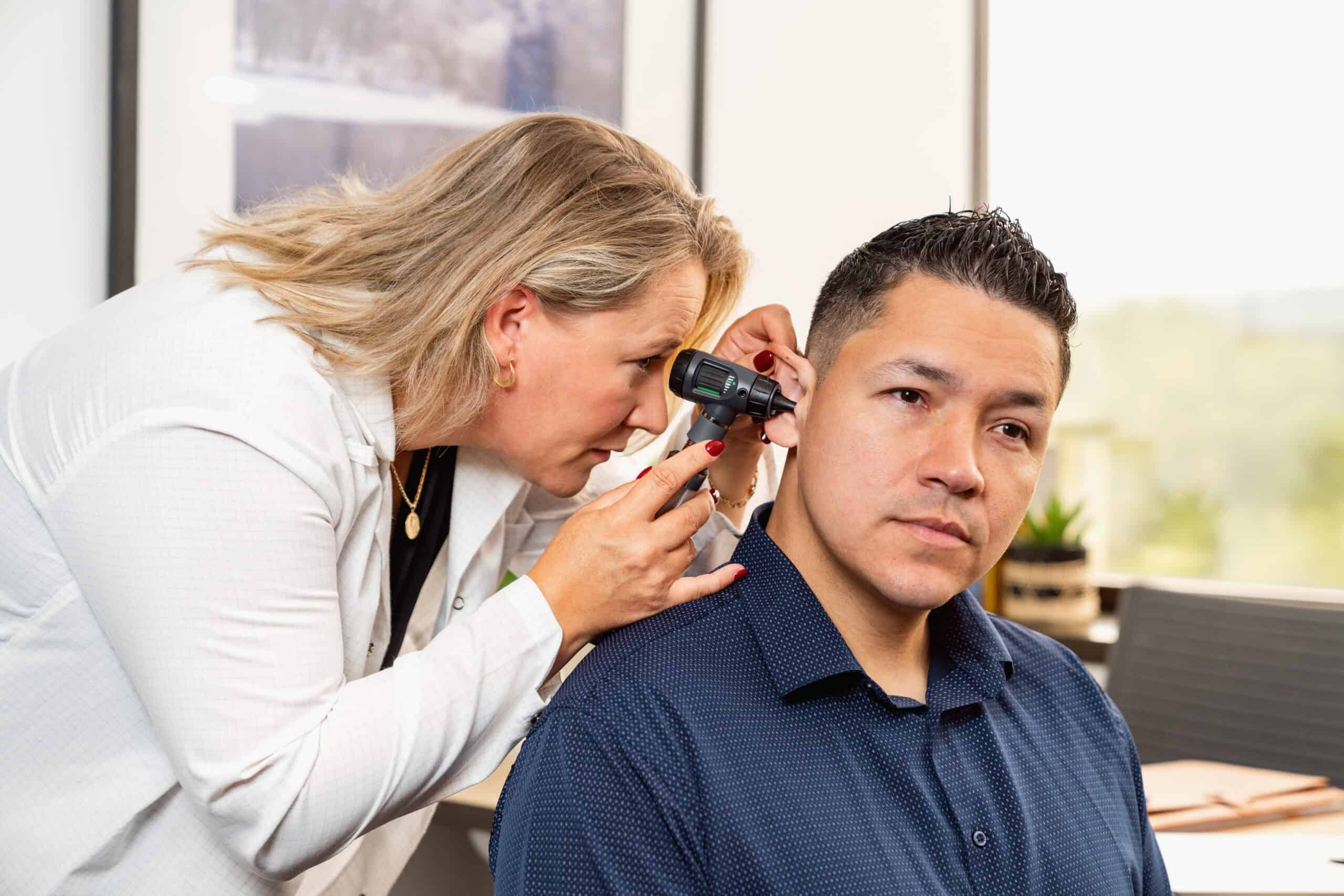 Female hearing aid specialist in white coat using an otoscope to examine male patient's ear canal. The specialist leans in with focused attention while the patient in navy blue shirt sits calmly. Bright, modern clinical office setting with framed artwork visible on walls in background. The ear examination is part of a comprehensive hearing evaluation.