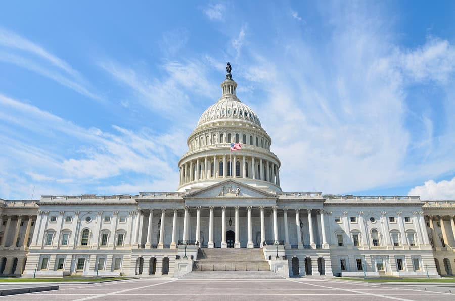 Front view of the United States Capitol building in Washington, D.C., featuring its iconic white dome topped with the Statue of Freedom. The neoclassical architecture shows rows of columns along the facade, grand entrance steps, and an American flag flying beneath the dome. Blue sky with wispy white clouds in the background.