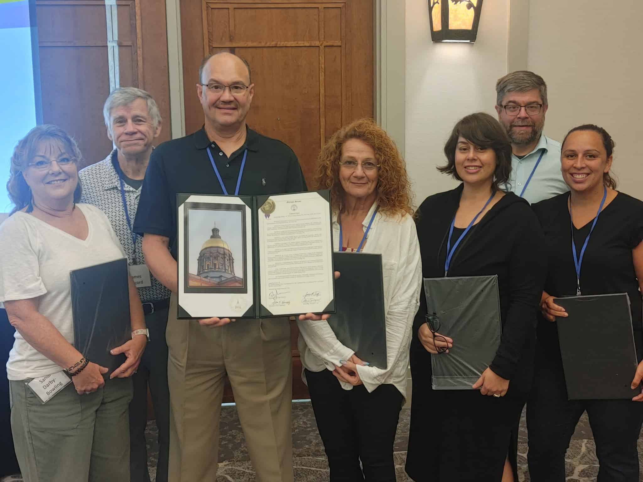 Group of seven people posing indoors for a formal photo. Two people in the center hold framed awards, including what appears to be a plaque with an image of a government building dome and an official certificate. The group members wear business casual attire and blue lanyards. A wooden door and wall sconce are visible in the background.