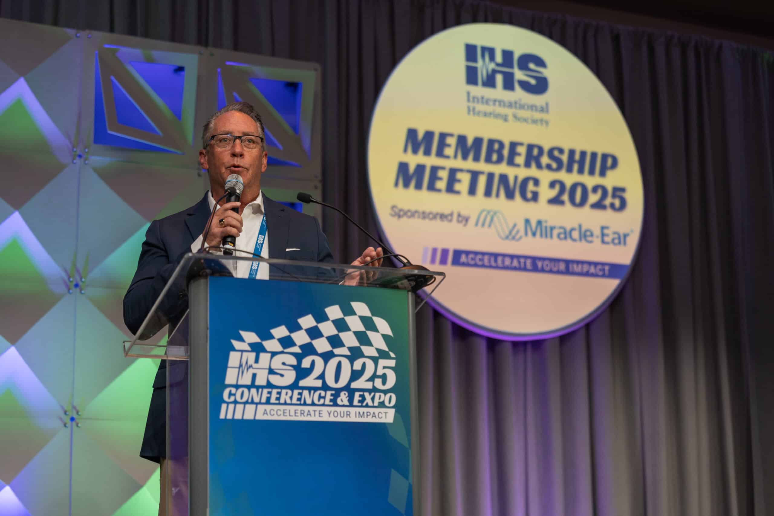 A speaker stands at a podium addressing an audience at the IHS 2025 Conference & Expo, with a large circular sign behind him reading “IHS International Hearing Society Membership Meeting 2025.”