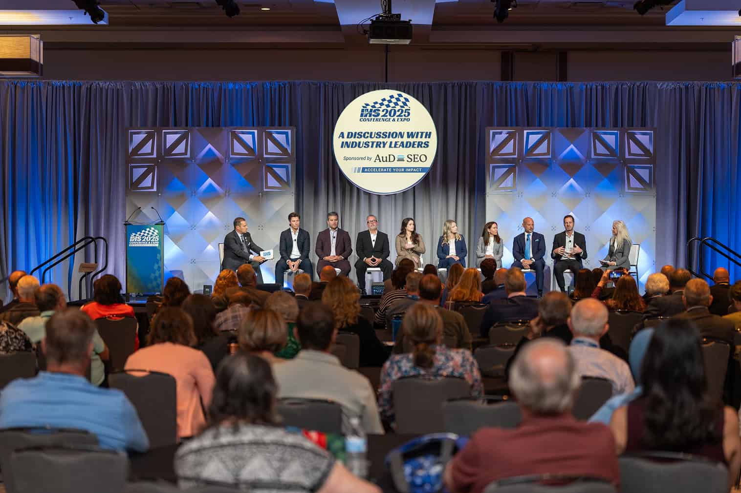 Industry leaders sit on stage for a panel discussion as a large audience listens at a conference.