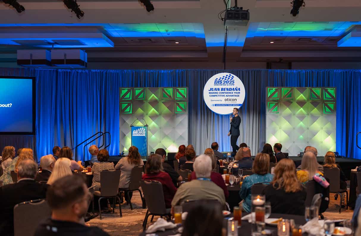 A keynote speaker stands on a stage addressing a large seated audience at a conference, with branded signage, colorful stage lighting, and attendees listening at round tables in a ballroom setting.