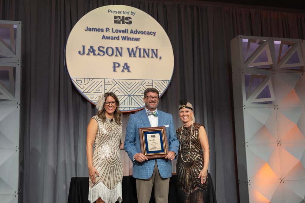 2025 James P. Lovell Advocacy Award Winner, Jason Winn, PA, accepting his award on stage with Executive Director, Alissa Parady, CAE, and President-Elect, Leanne Polhill, BA, BC-HIS