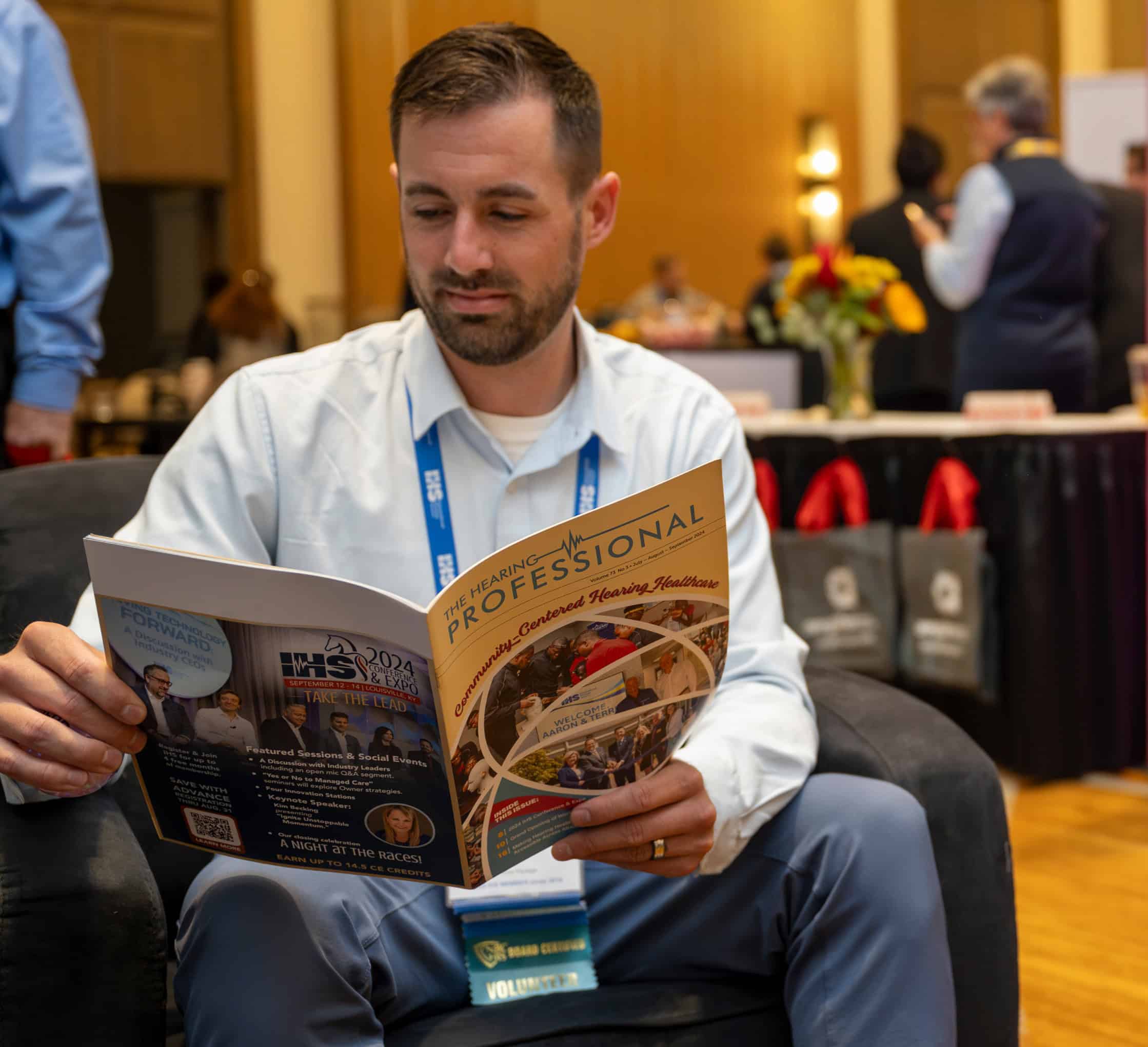 A man wearing a conference badge sits in a lounge chair reading a professional magazine at a conference expo, with exhibitor booths and attendees visible in the background.