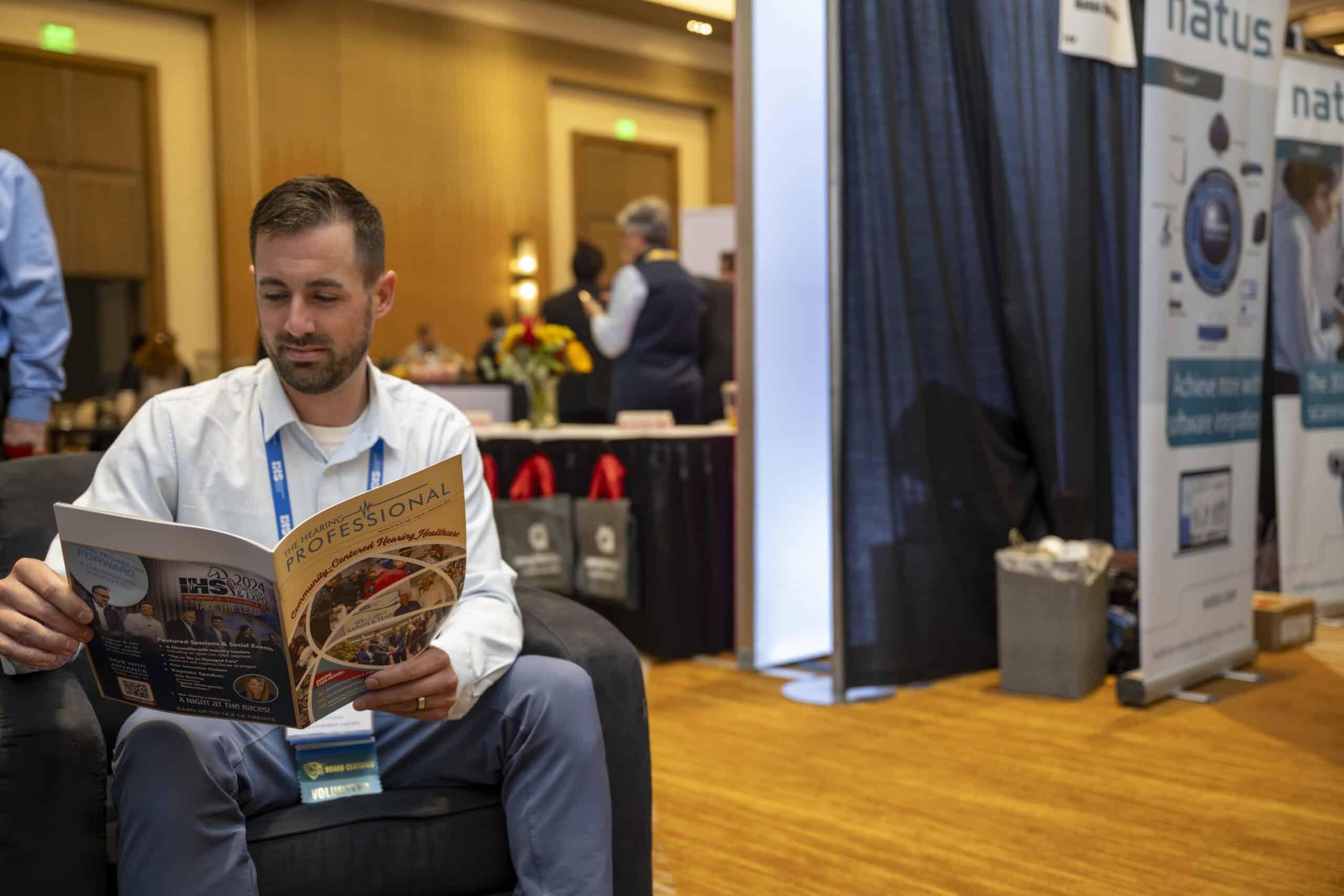 Conference attendee sits in a chair reading The Hearing Professional magazine.