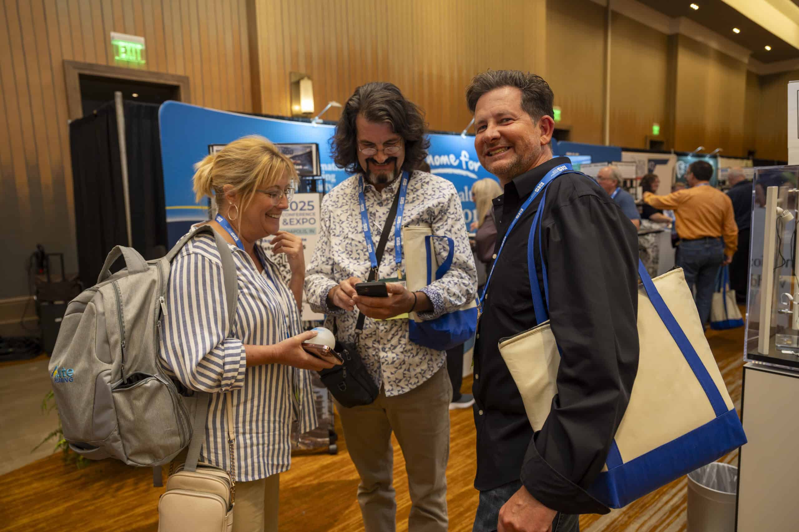Three conference attendees stand together at an expo hall, smiling and chatting while looking at a phone, wearing event badges and carrying tote bags with exhibitor booths visible behind them.