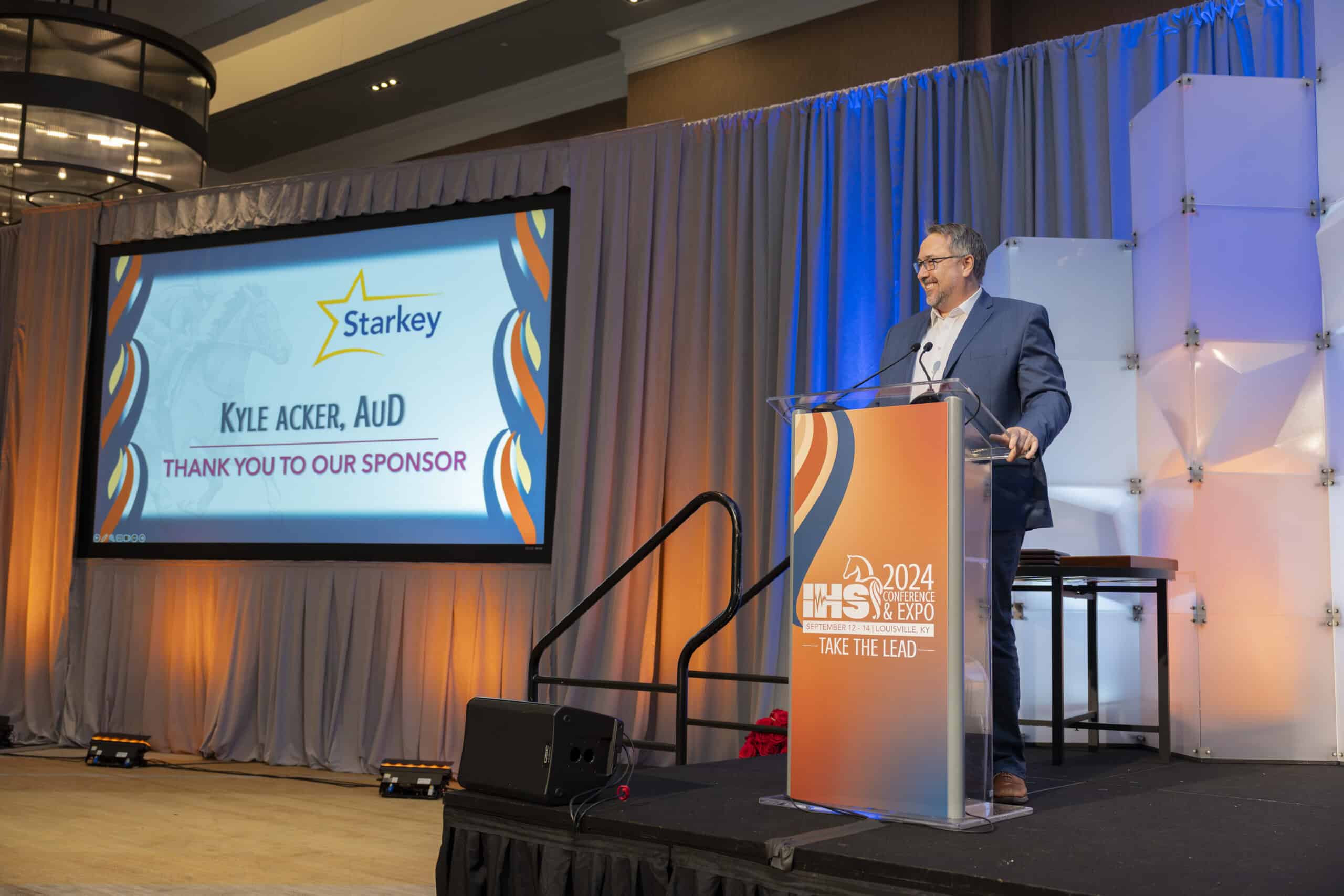 Kyle Acker, AuD pictured on stage during the IHS Conference & Expo where Starkey is noted as a sponsor.