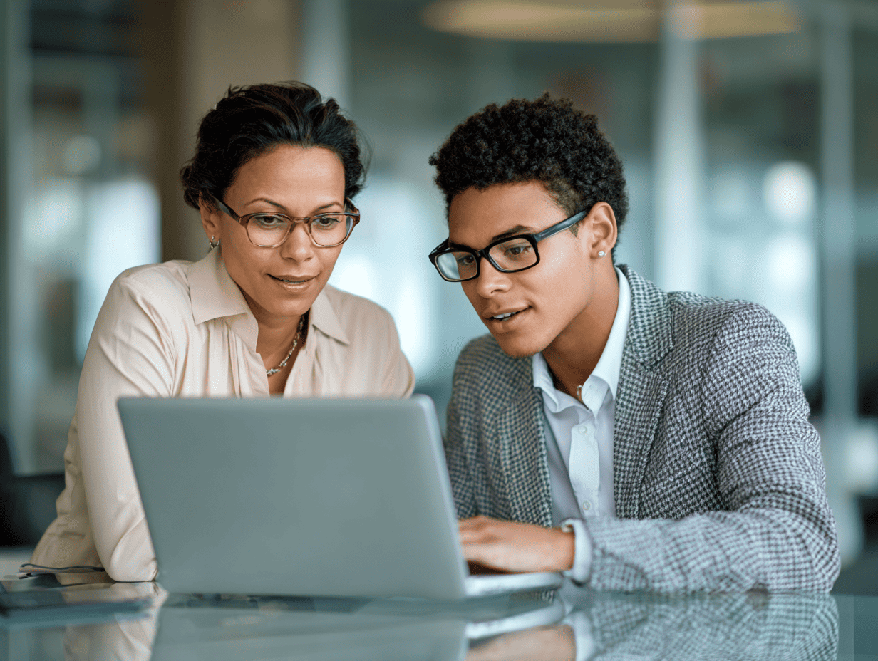Two people wearing glasses sit side by side at a table, focused on a laptop screen as they work together in a bright, modern office with glass walls in the background.