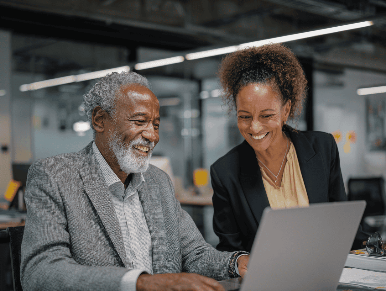An older man with gray hair and a beard smiles while using a laptop at a desk, as a woman beside him leans in and smiles; they appear to be collaborating in a modern office with glass walls and overhead lighting.