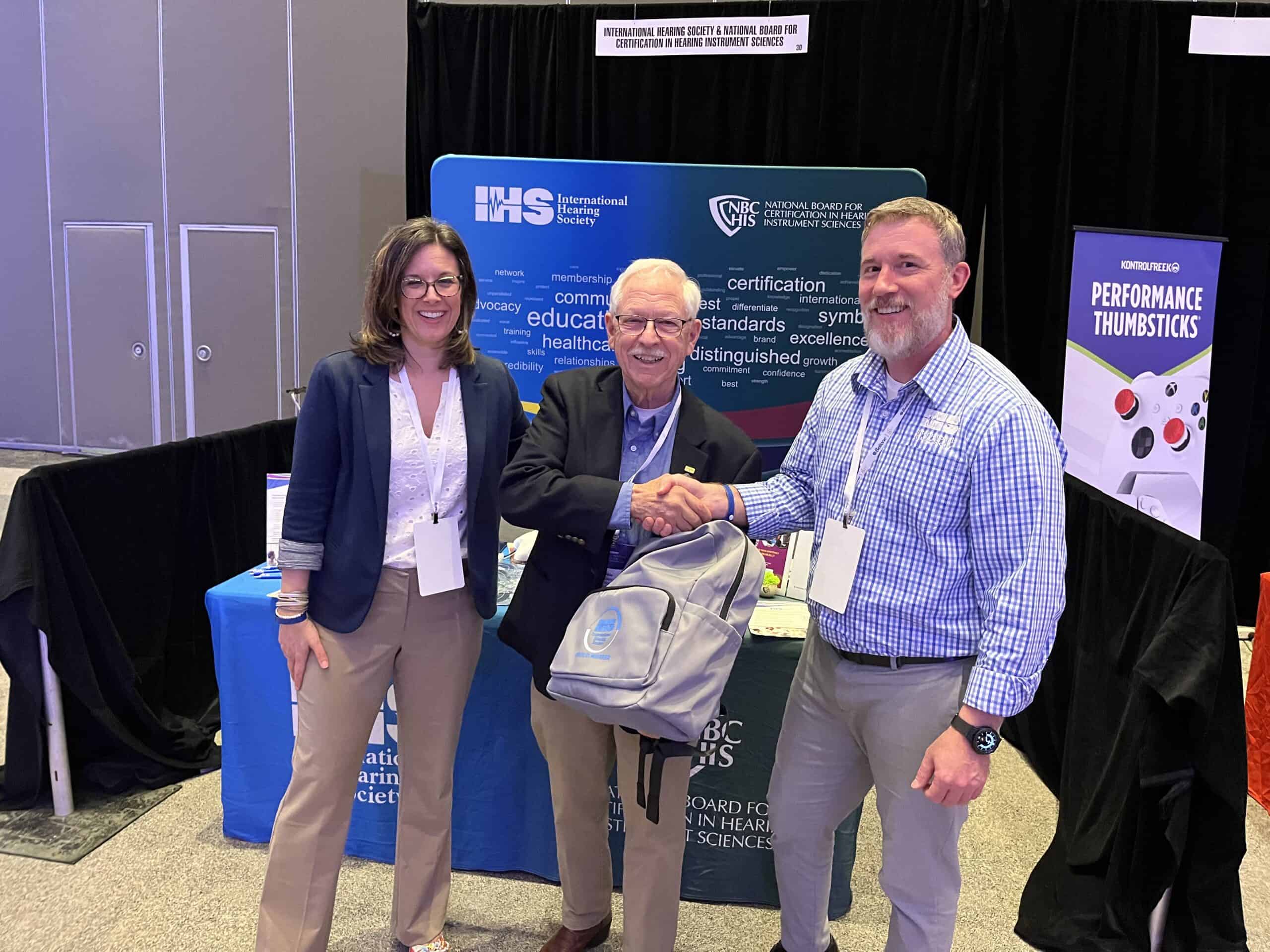Three conference attendees at an IHS booth, smiling with IHS merch.