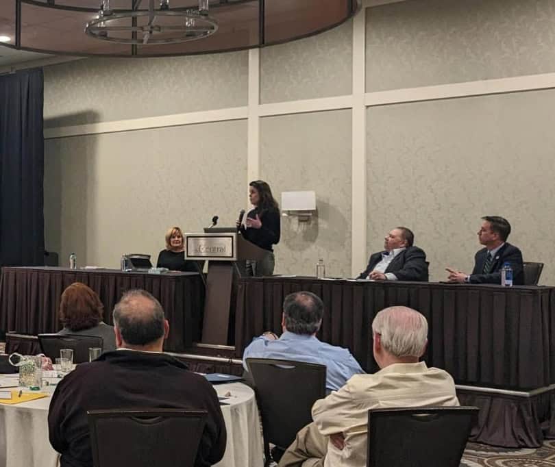 A woman stands at a podium speaking to an audience in a conference room, with several panelists seated at a long table behind her and attendees listening at round tables in the foreground.
