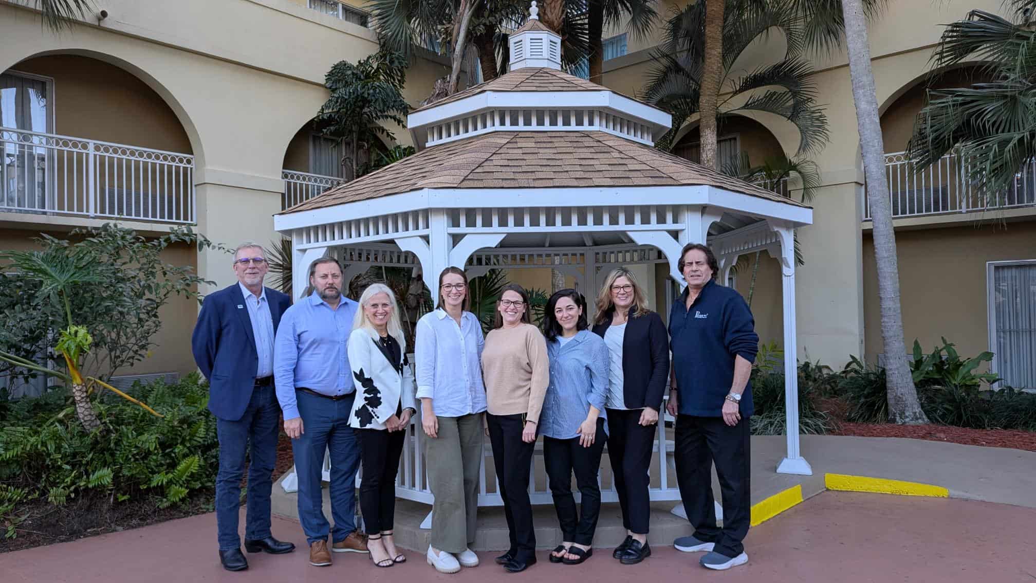 Group of nine adults stand together outdoors in front of a white gazebo at a conference venue.
