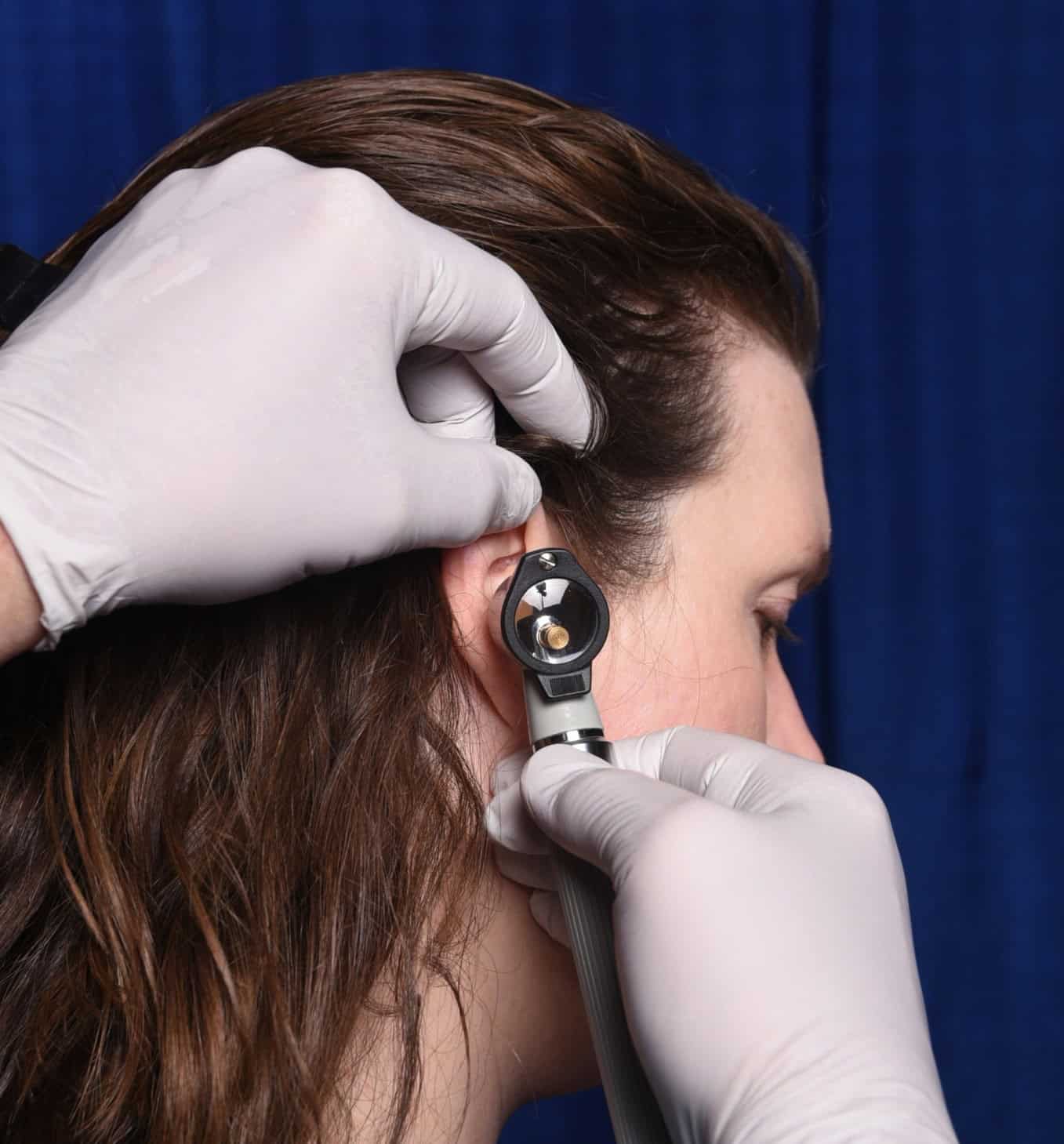Instructor demonstrates bracing for otoscopy. Focus is on the instructor's hands and the person's side of their face. Otoscope handle is pointing down aligned with the instructor's wrist. The instructor's hand without the instrument is holding the top of the ear while the fingers of the instrument hand brace against the cheek.