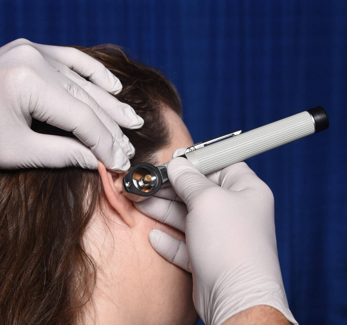 Instructor demonstrates bracing for otoscopy. Focus is on the instructor's hands and the person's side of their face. Otoscope handle is pointing toward the top left corner of the image. The fingers of the instrument hand brace against the cheek, while the fingers of the non-instrument hand both brace against the head and also pull the pinna up for viewing.