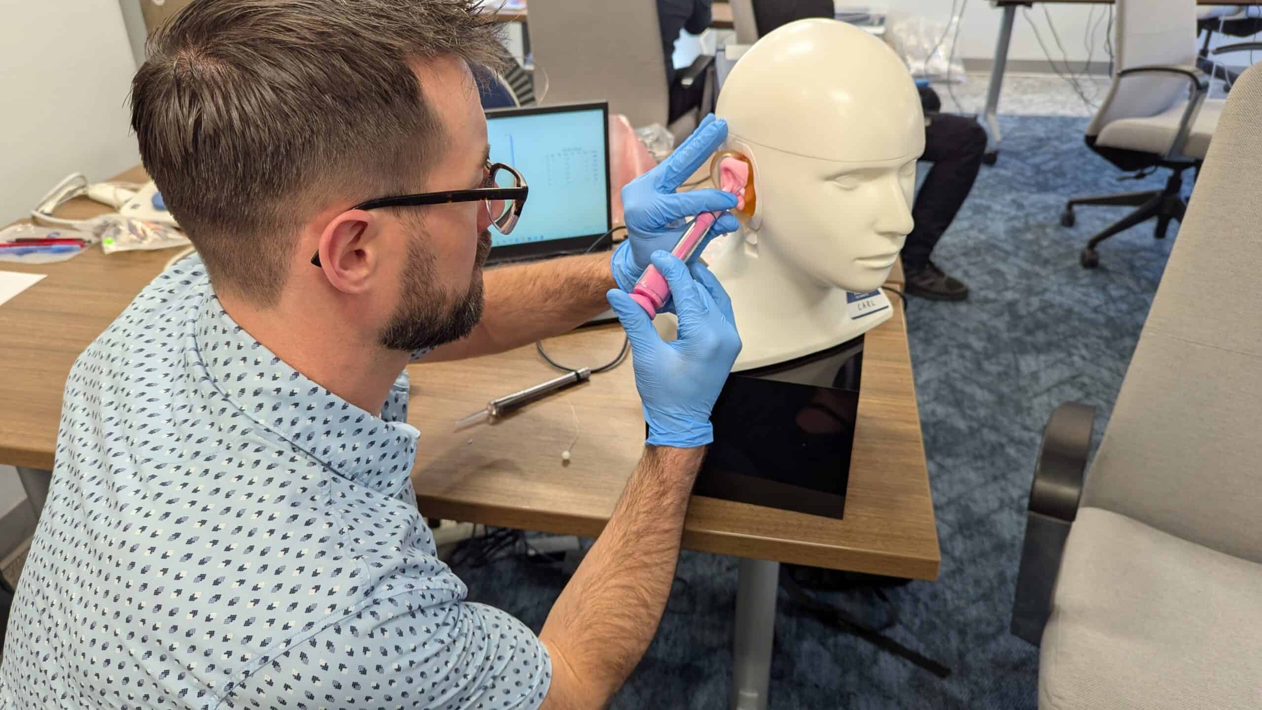 A man wearing gloves carefully works on a mannequin head at a desk, inserting or adjusting an ear mold using a handheld tool, with a computer and training materials visible in an office or classroom setting.
