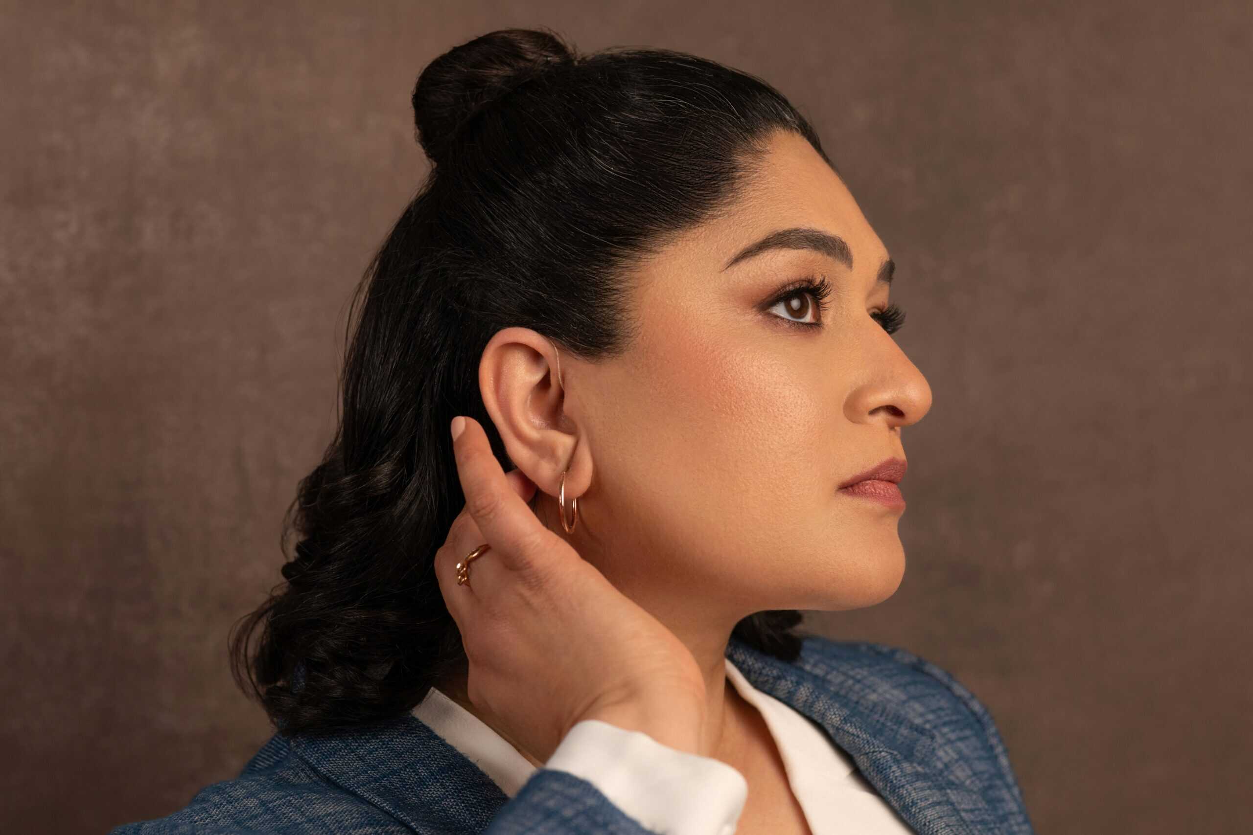 Profile view of young woman with dark hair styled in half-up bun, wearing modern discreet hearing aid. She touches her ear gently while gazing forward confidently, dressed in blue blazer over white shirt with gold hoop earrings. Neutral brown background emphasizes the natural, stylish appearance of contemporary hearing aid technology.