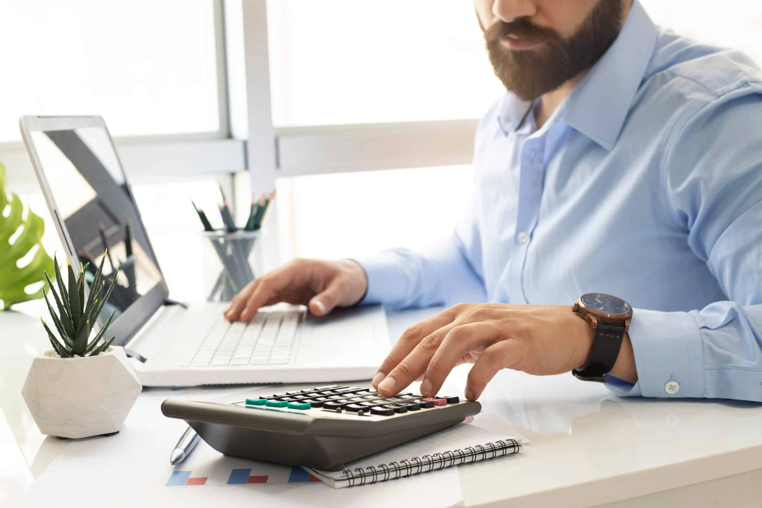 businessman running some calculations on a desk calculator with their laptop.