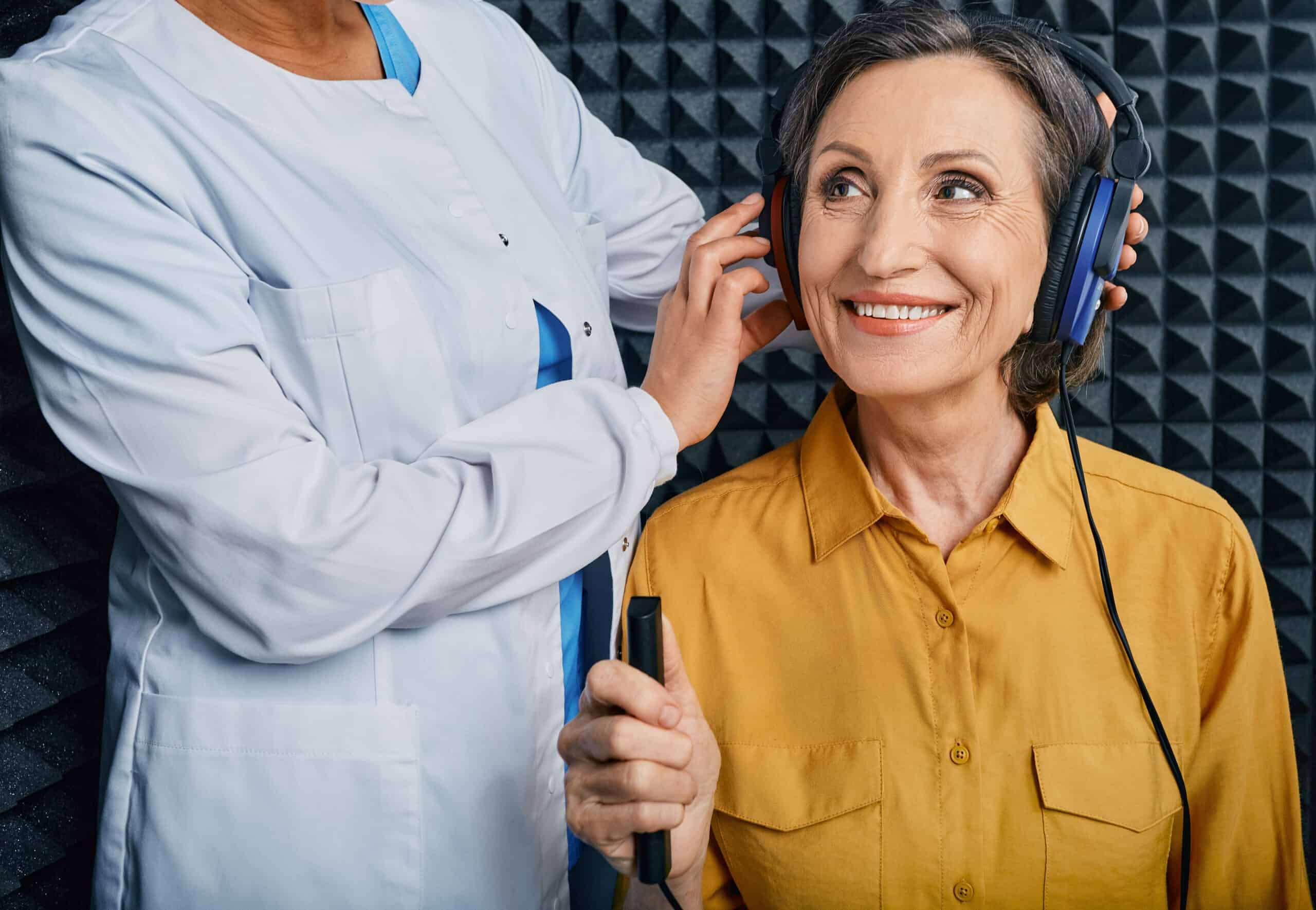 Hearing aid specialist in white coat placing professional headphones on smiling senior woman in yellow shirt during hearing assessment. The patient looks upward with an engaged, cheerful expression while holding response button. Sound-dampening acoustic foam wall panels visible in background of audiology testing booth.