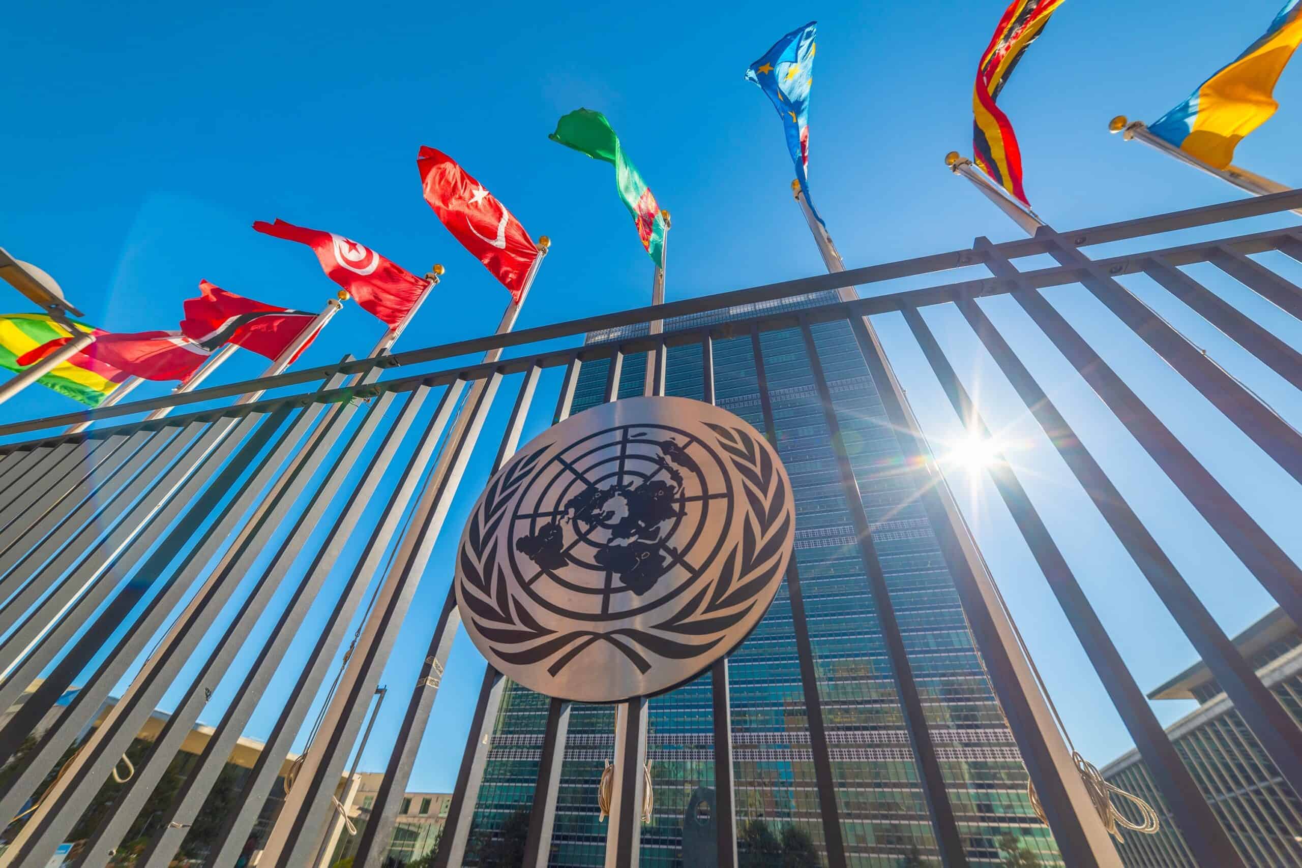View looking up at UN headquarters building with prominent United Nations emblem on metal gate in foreground. Multiple colorful international flags wave against clear blue sky above the building. Sunburst visible at right side of modern glass and steel structure.