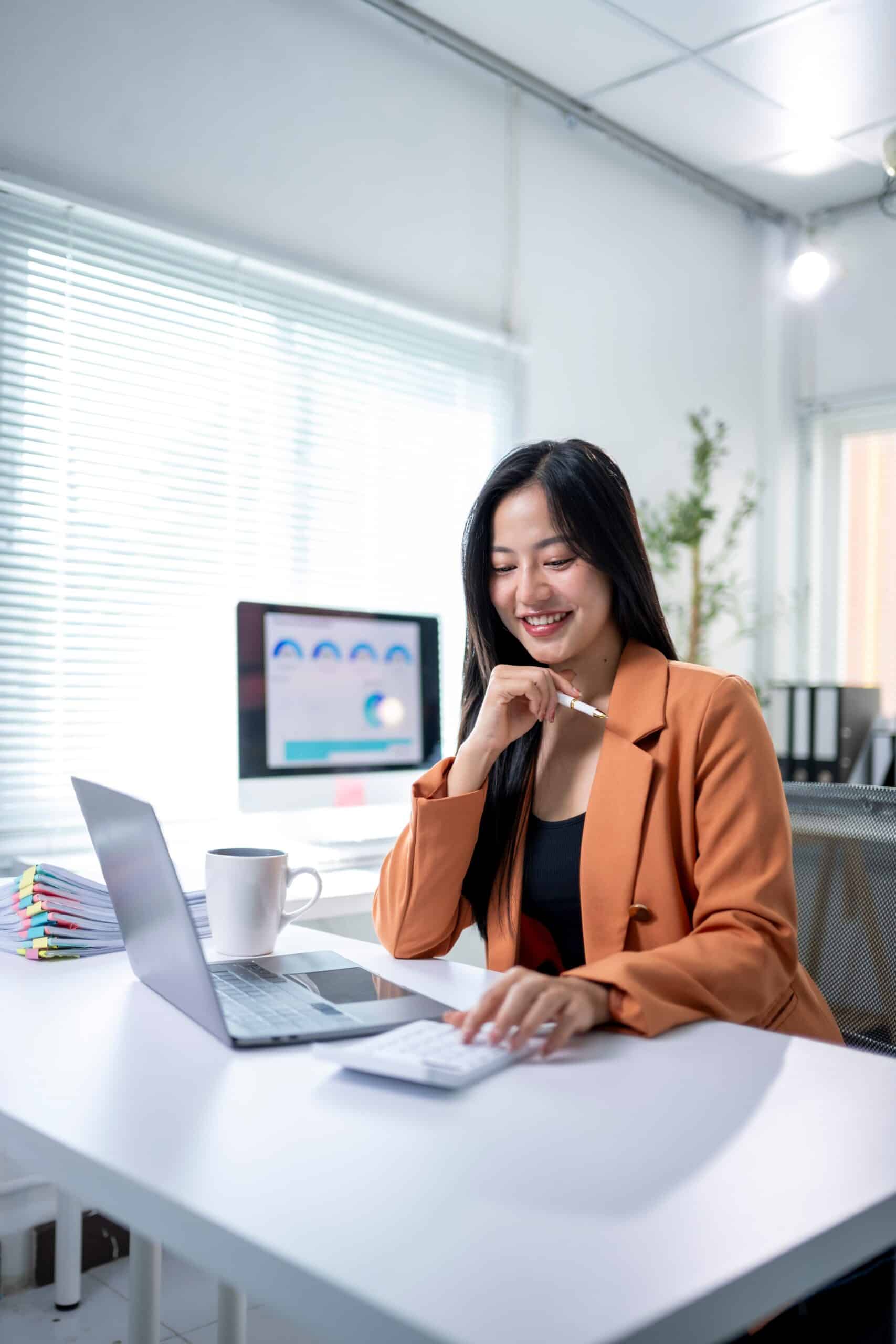 Woman smiling while working at a desk with a laptop and calculator in a bright office.