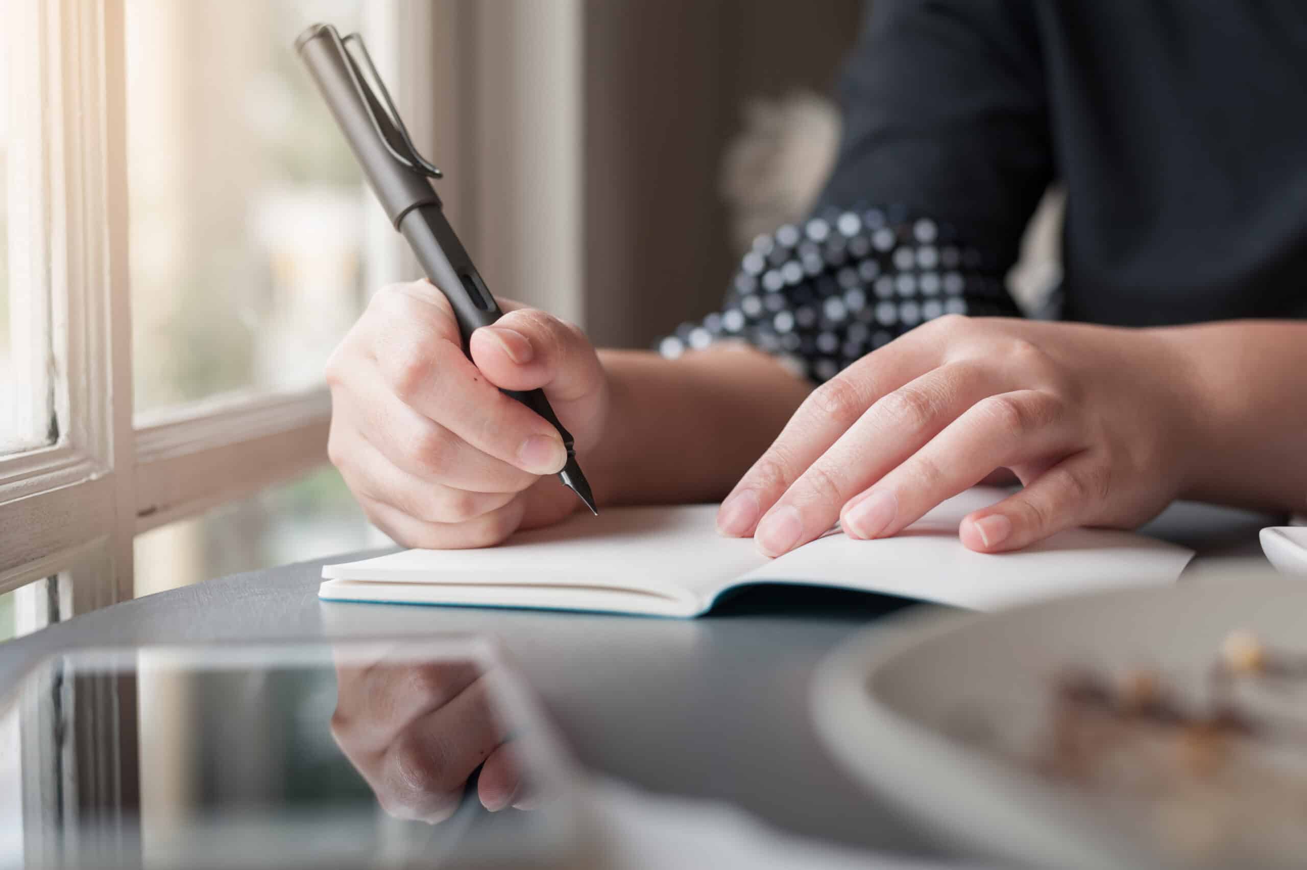 A close-up of a person writing in a small notebook with a pen at a table near a window.