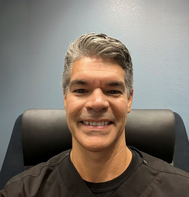 Man with graying hair and a black scrub top, smiles at the camera.